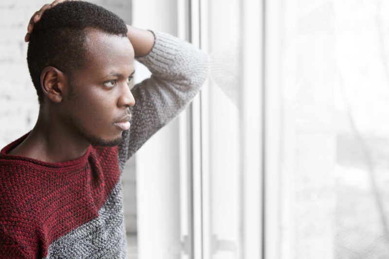 Sad dark-skinned male in casual sweater touching his head and staring through window glass with unhappy and depressed look, suffering from creative frustration, can't find inspiration at new job