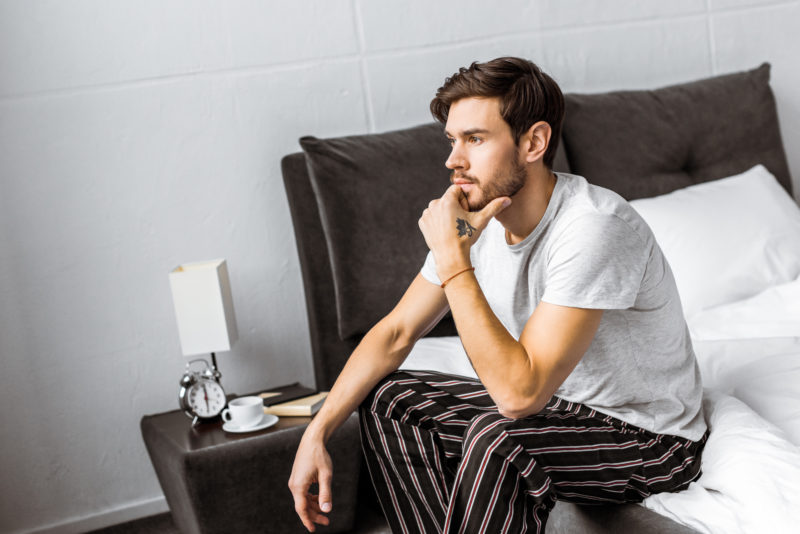 thoughtful young man in pajamas sitting on bed and looking away in the morning
